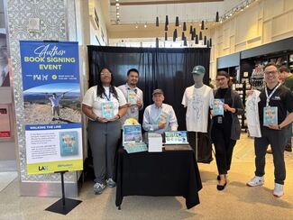 Several travelers stand in front of a bookstore, smiling and posing with the author sitting at a table.